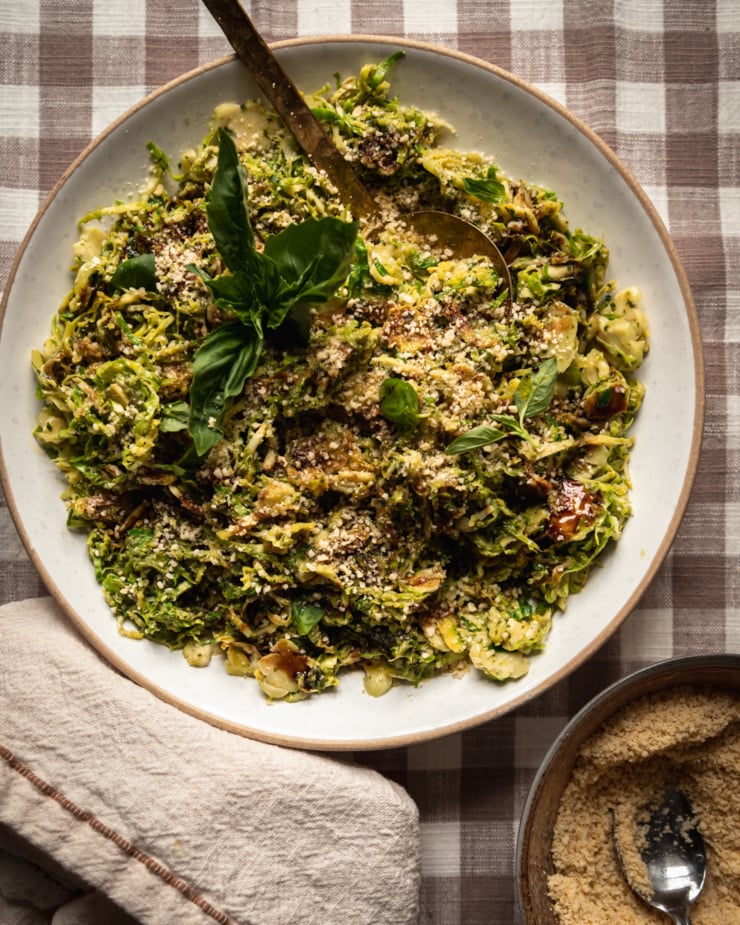 An overhead shot of sautéed shaved brussels sprouts in a shallow bowl, topped with balsamic glaze, fresh basil and vegan "parmesan"
