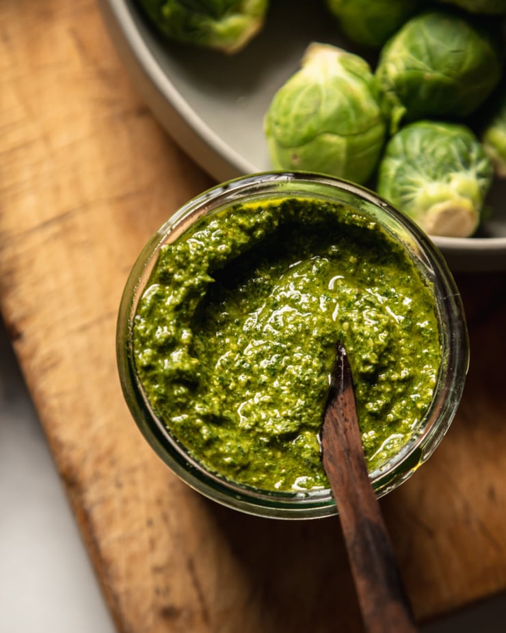 An overhead shot shows a jar of vegan pesto with a wooden spoon sticking out.