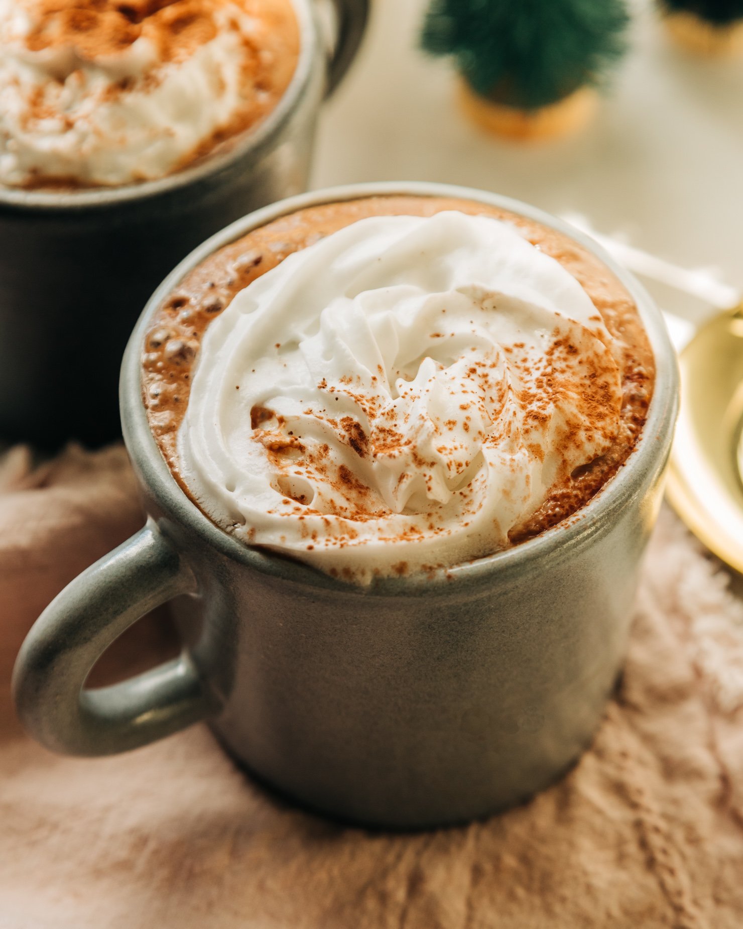 A 3/4 angle shot of vegan almond butter hot chocolate in a mug, topped with a swirl of coconut whipped cream and a sprinkle of cocoa powder.