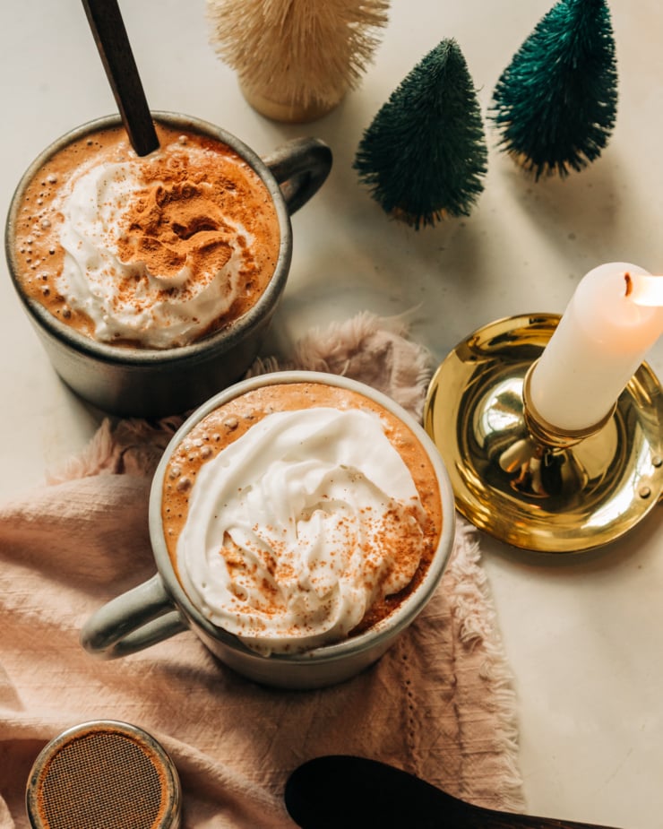 An overhead shot of 2 mugs of vegan almond butter hot chocolate in a mug, topped with a swirl of coconut whipped cream and a sprinkle of cocoa powder. A lit candle in a brass holder is nearby.