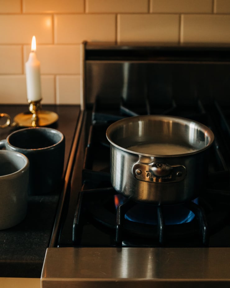 A 3/4 angle shot of a saucepan of non-dairy milk heating up on a gas stove. A lit candle is nearby.