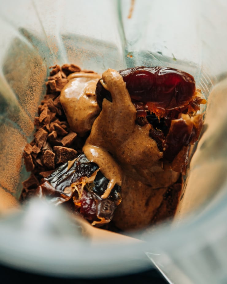 An overhead shot of almond butter, chopped chocolate, dates, and cocoa powder in a blender pitcher.