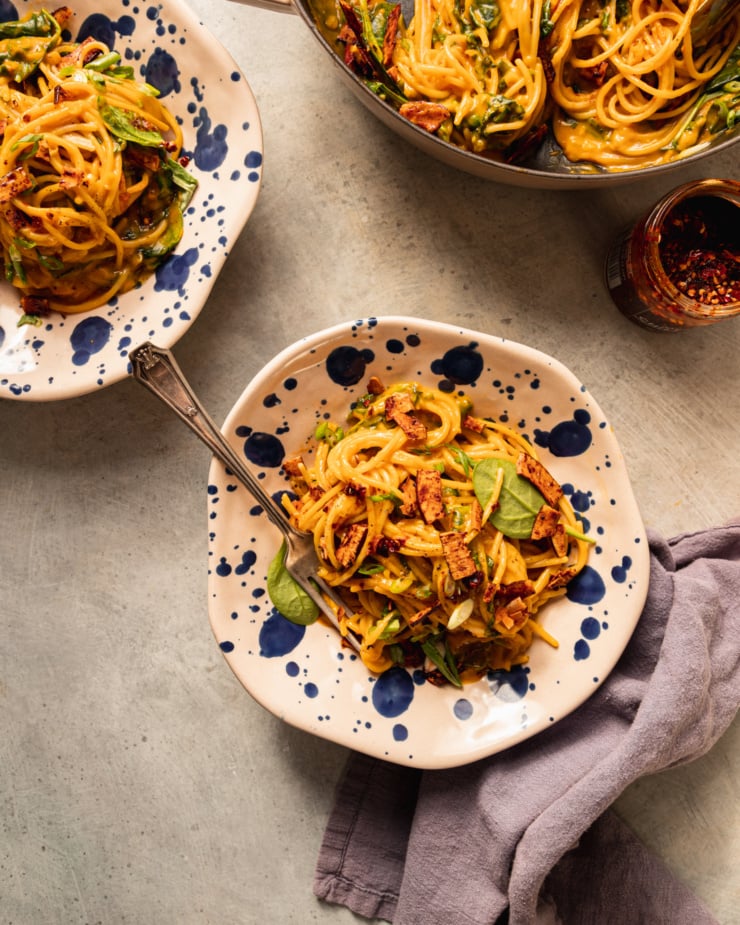 An overhead shot shows two servings of butternut coconut noodles in wide bowls along with the braiser serving pot that also contains the noodles.