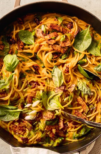 An up close, overhead shot shows a braiser pot filled with creamy butternut coconut noodles mixed with fresh basil, green onions, and crunchy bits of smoky coconut on top.