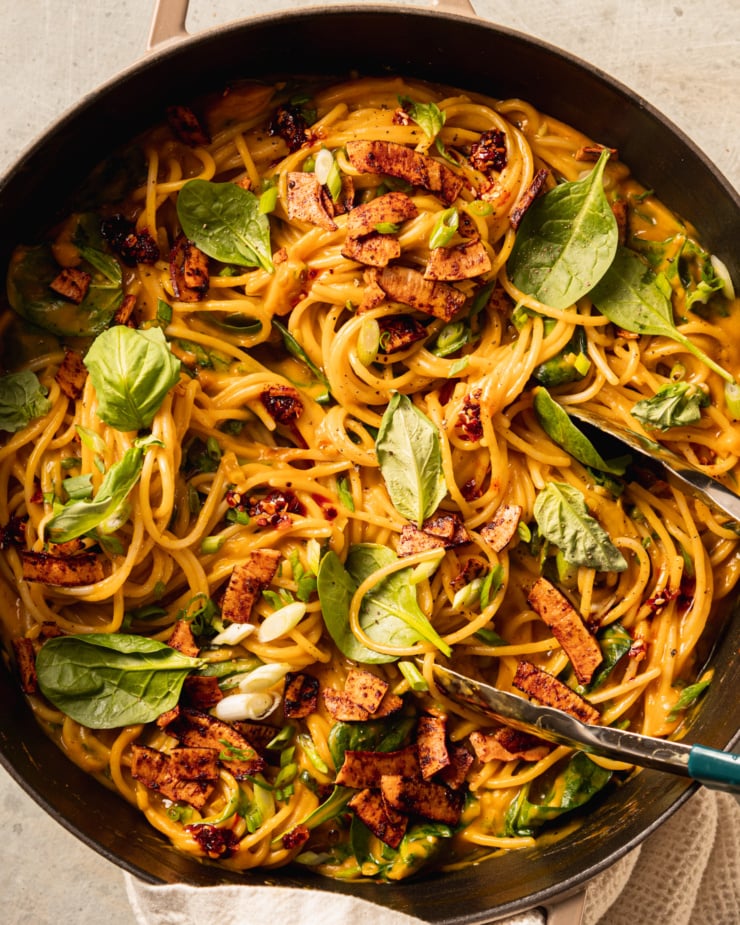 An up close, overhead shot shows a braiser pot filled with creamy butternut coconut noodles mixed with fresh basil, green onions, and crunchy bits of smoky coconut on top.