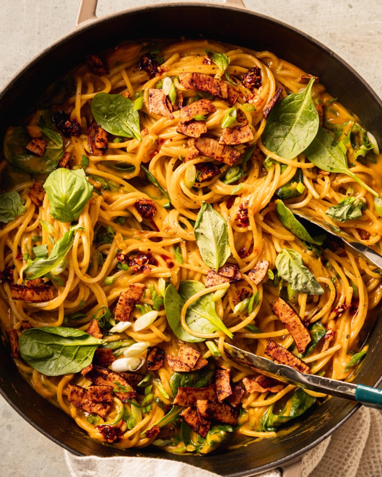 An up close, overhead shot shows a braiser pot filled with creamy butternut coconut noodles mixed with fresh basil, green onions, and crunchy bits of smoky coconut on top.