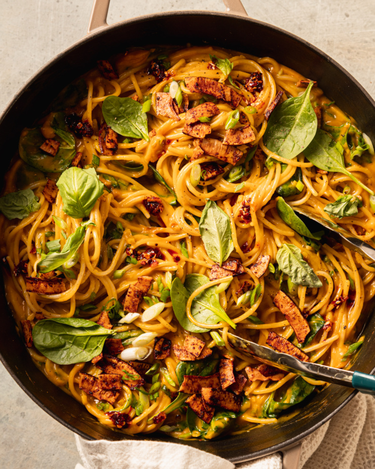 An up close, overhead shot shows a braiser pot filled with creamy butternut coconut noodles mixed with fresh basil, green onions, and crunchy bits of smoky coconut on top.