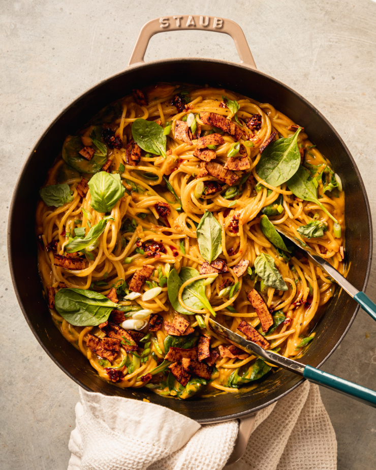 An overhead shot shows a braiser pot filled with creamy butternut coconut noodles mixed with fresh basil, green onions, and crunchy bits of smoky coconut on top.