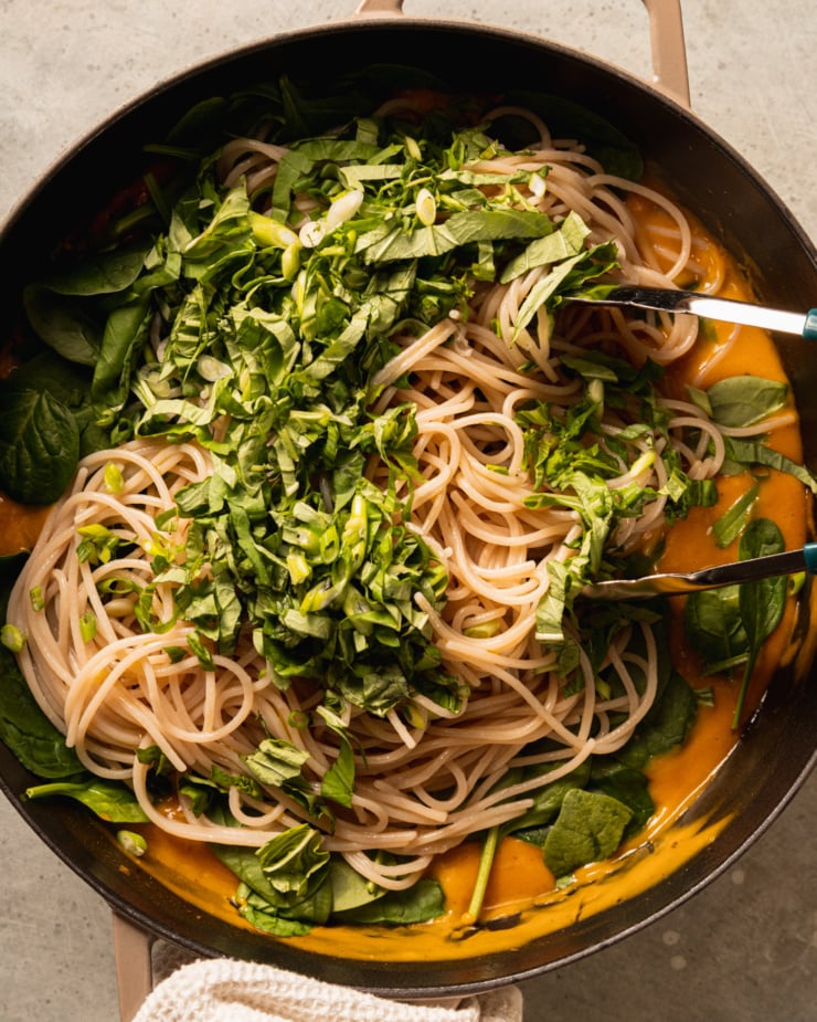 An overhead shot shows a braiser filled with a creamy butternut squash sauce, cooked rice noodles, baby spinach, chopped basil, and green onions.