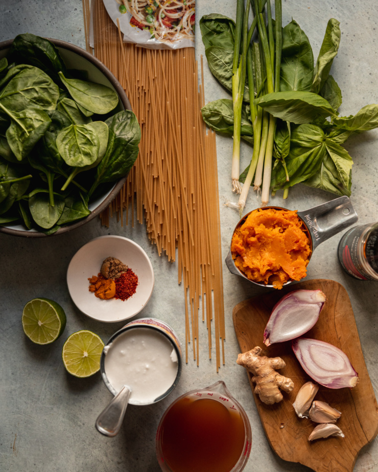 An overhead shot shows ingredients for a vegan butternut squash noodle dish with herbs, spices, and greens.