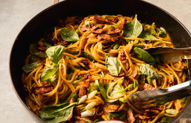 An up close, slight 3/4 angle shot shows a braiser pot filled with creamy butternut coconut noodles mixed with fresh basil, green onions, and crunchy bits of smoky coconut on top.