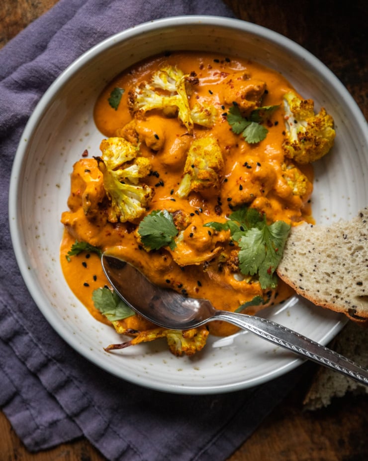 An overhead shot of a single serving of a saucy cauliflower dish that is garnished with cilantro and nigella seeds in a wide bowl. There is a blue linen napkin nearby.