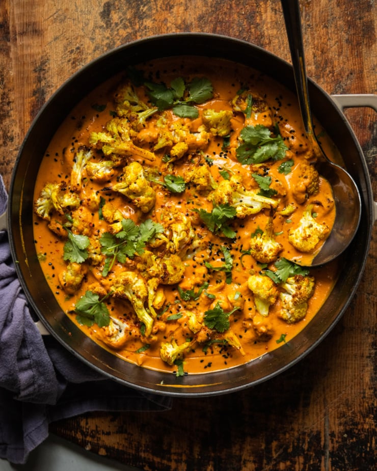 An overhead shot of roasted cauliflower in a coconut tomato sauce. The dish is garnished with fresh cilantro, nigella seeds, and ground chillies.