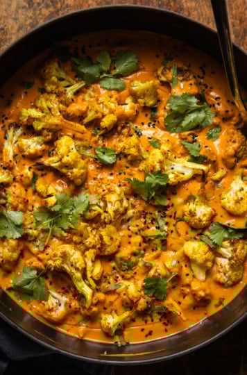 An overhead shot of roasted cauliflower in a coconut tomato sauce. The dish is garnished with fresh cilantro, nigella seeds, and ground chillies.
