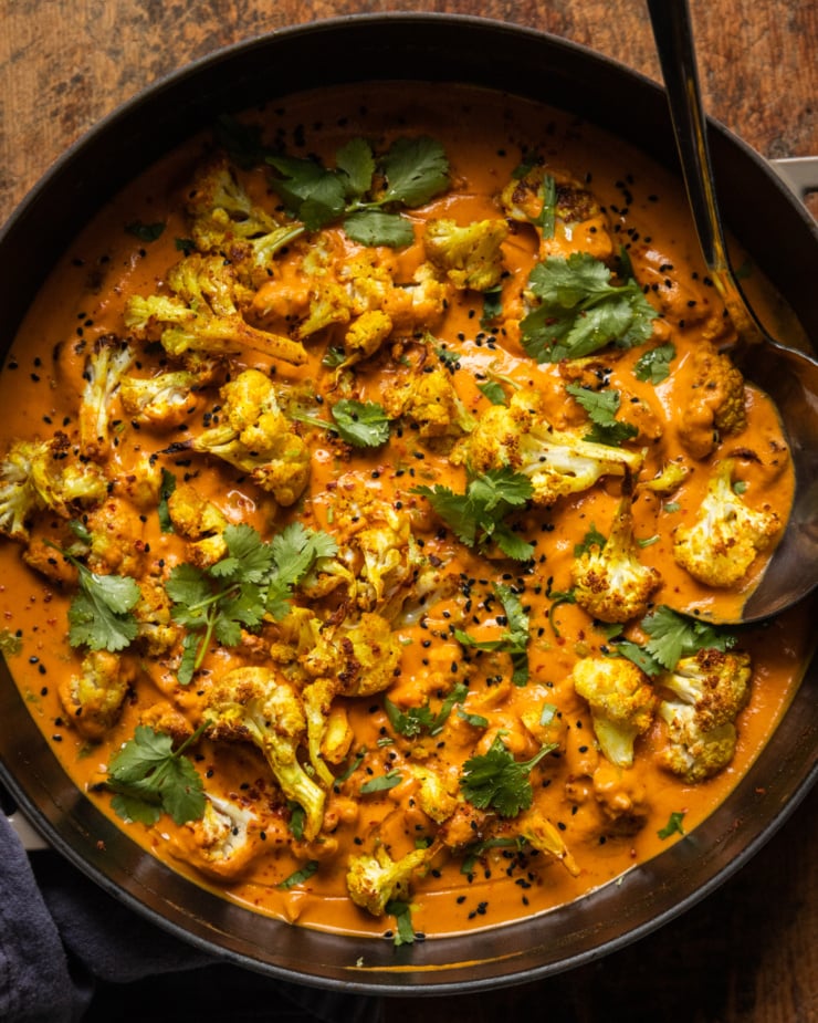 An overhead shot of roasted cauliflower in a coconut tomato sauce. The dish is garnished with fresh cilantro, nigella seeds, and ground chillies.