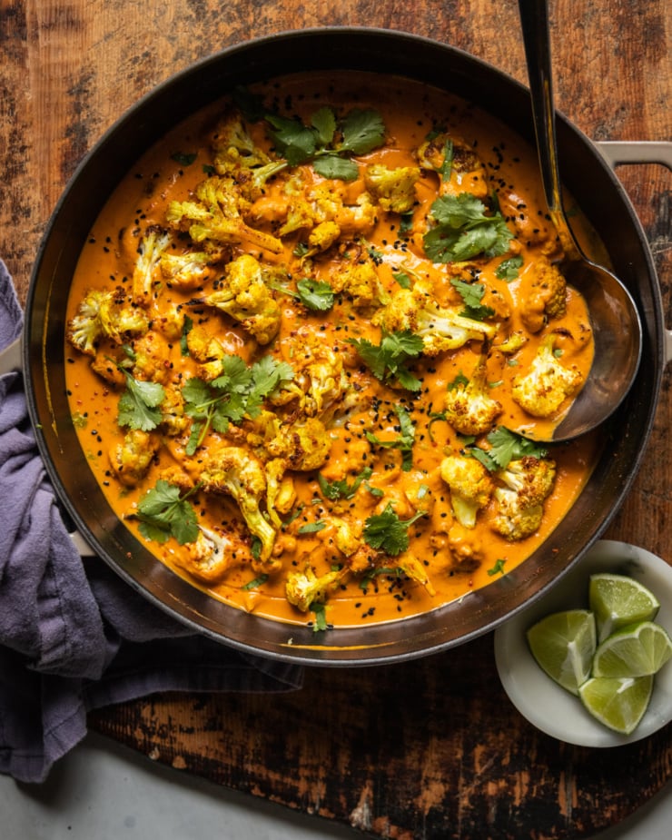 An overhead shot of roasted cauliflower in a coconut tomato sauce. The dish is garnished with fresh cilantro, nigella seeds, and ground chillies.