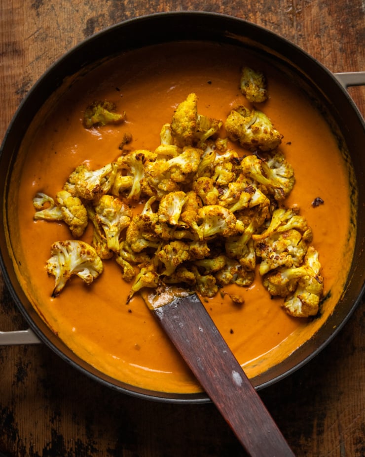 An overhead shot shows roasted cauliflower florets that have just been added to a pot of silky and rich coconut tomato sauce.