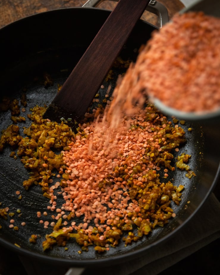A slight 3/4 angle shot shows split red lentils being added to a pot of sautรฉed shallots and spices.