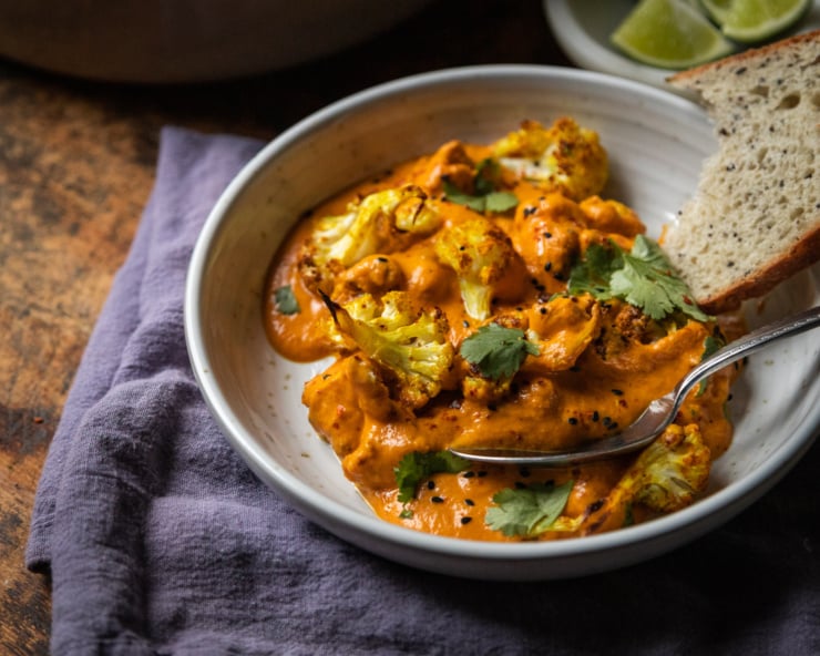 A 3/4 angle shot of a single serving of a saucy cauliflower dish that is garnished with cilantro and nigella seeds in a wide bowl. There is a blue linen napkin nearby.