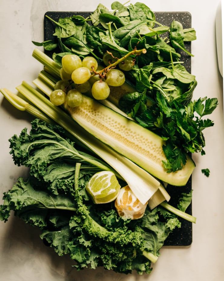 An overhead shot shows a black cutting board loaded up with green juice ingredients: kale, peeled lemon, peeled lime, celery, a halved cucumber, green grapes, baby spinach, and flat leaf parsley.