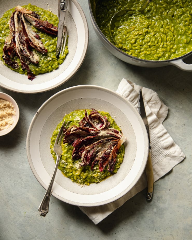 An overhead shot of two servings of broccoli rabe risotto in wide, light grey and white bowls. The risotto is topped with roasted wedges of radicchio and vegan "parmesan." The pot of deep green risotto is nearby.