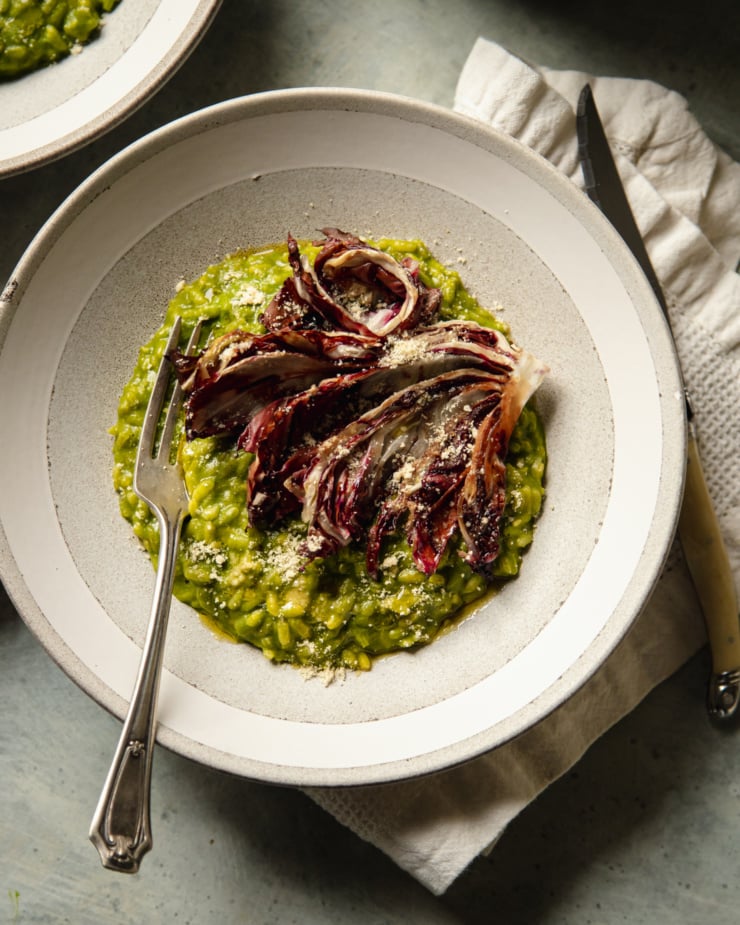 An overhead shot of a serving of broccoli rabe risotto in a wide light grey and white bowl. The risotto is topped with a roasted wedge of radicchio and vegan "parmesan"