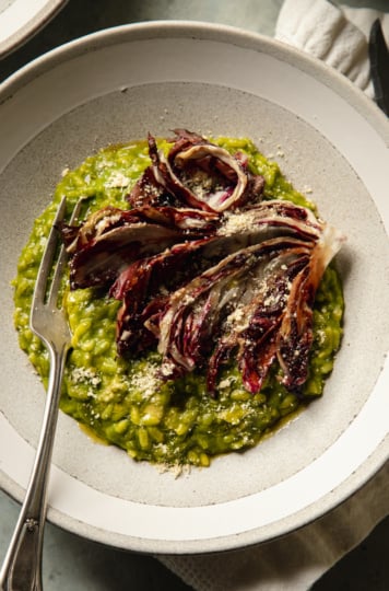 An overhead shot of a serving of broccoli rabe risotto in a wide light grey and white bowl. The risotto is topped with a roasted wedge of radicchio and vegan "parmesan"