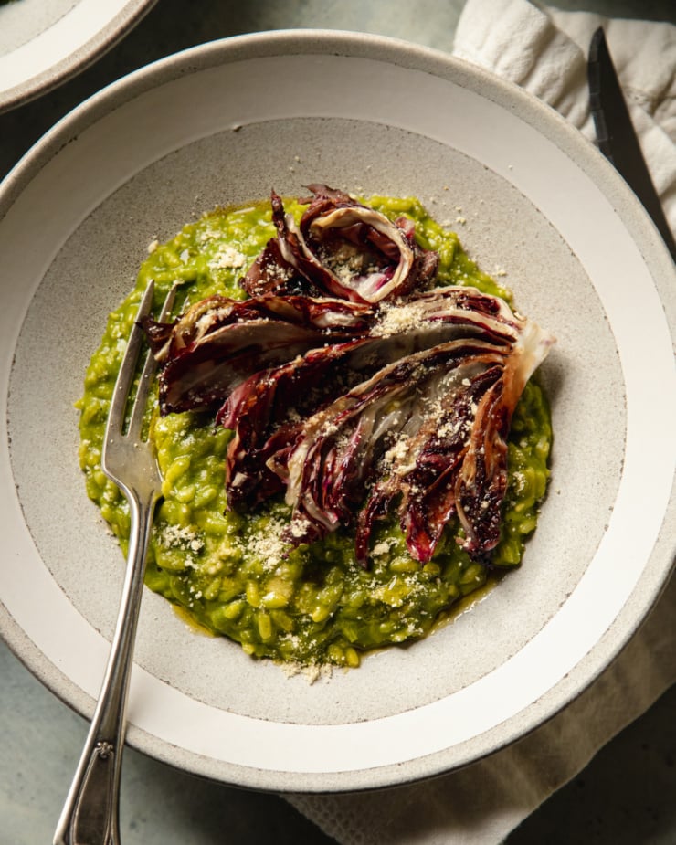 An overhead shot of a serving of broccoli rabe risotto in a wide light grey and white bowl. The risotto is topped with a roasted wedge of radicchio and vegan "parmesan"