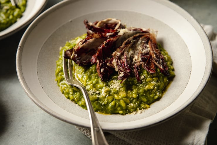 An up close, 3/4 angle shot shows a shallow grey bowl with a serving of deep green broccoli rabe risotto topped with a roasted wedge of radicchio.