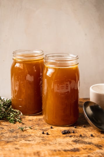 A head-on shot shows two jars of vegetable stock on a rough wooden cutting board. Sprigs of thyme, black peppercorns, a ladle, and a pot of salt are seen to the side.