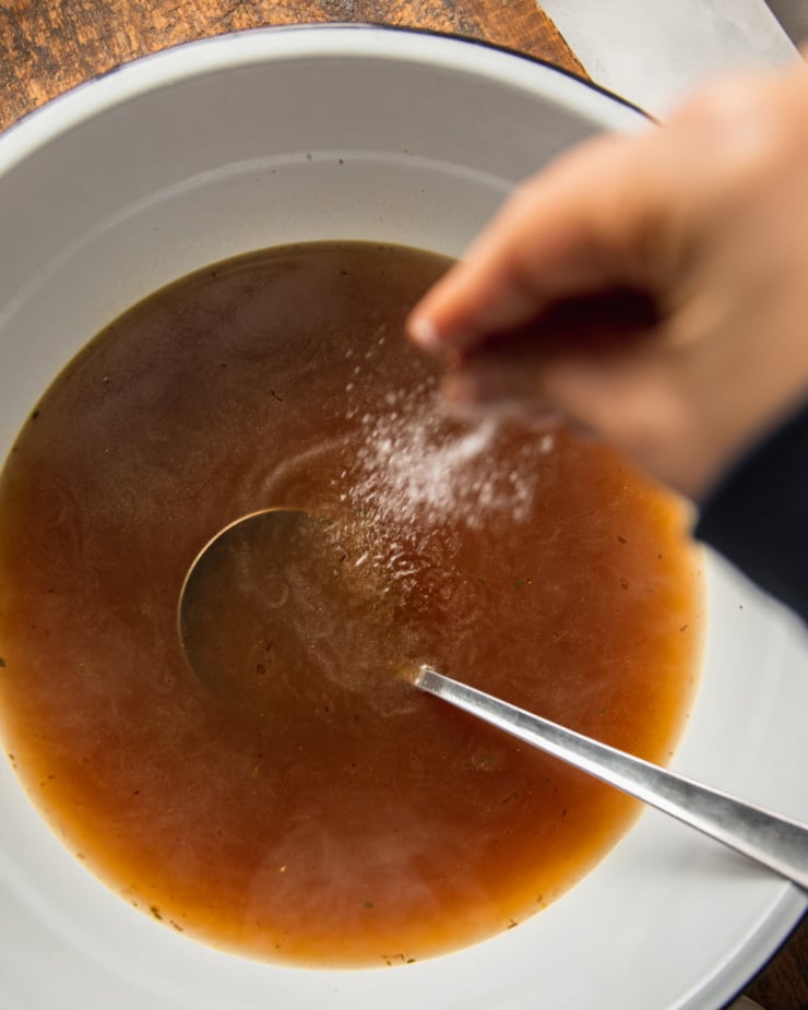 An overhead image shows a hand liberally seasoning a bowl of vegetable stock with a ladle sticking out.