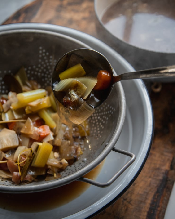 A 3/4 angle shot shows a ladle transferring vegetables and stock to a strainer and bowl setup.