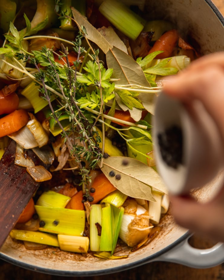 An overhead shot shows. hand sprinkling some peppercorns into a pot filled with sautéed vegetable stock ingredients.