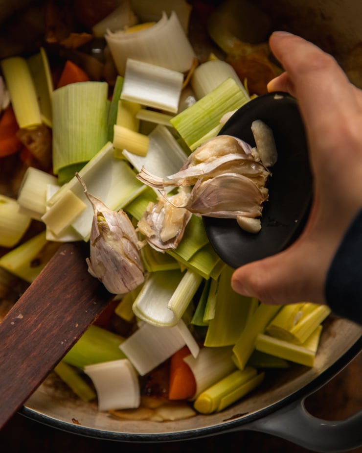 An overhead shot shows a hand lowering a small bowl of smashed garlic cloves into a pot of sautéed onions, carrots, celery, and chopped leeks.