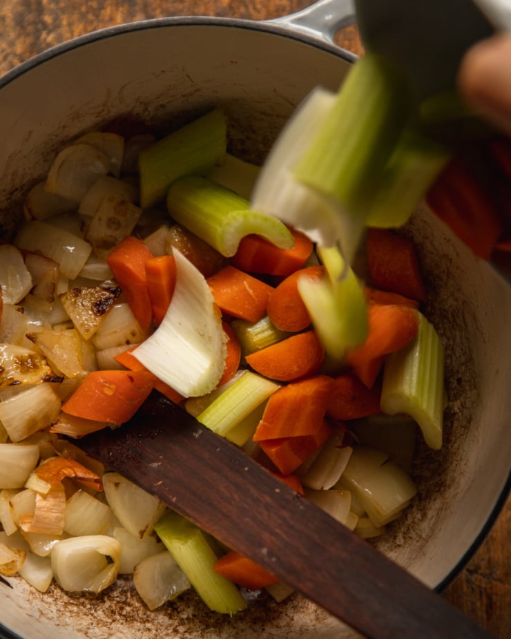 An overhead shot shows chopped carrots and celery being added to a pot of sautéed onions.