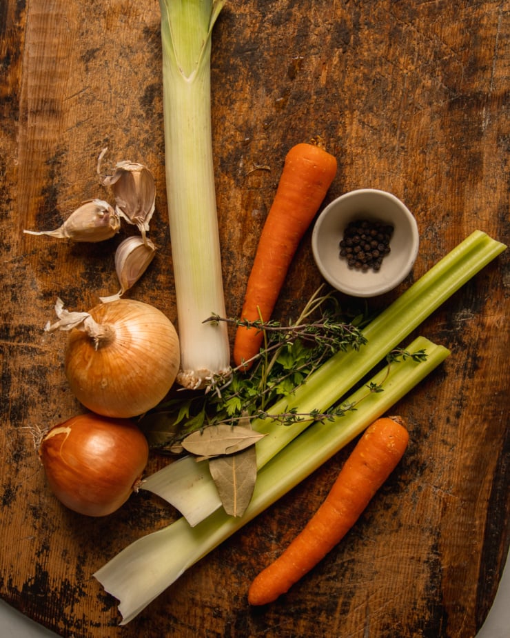 An overhead shot shows ingredients used in vegetable stock: a leek, carrots, celery, onions, garlic, bay leaves, thyme, parsley, and black peppercorns; all on a rough wooden cutting board.