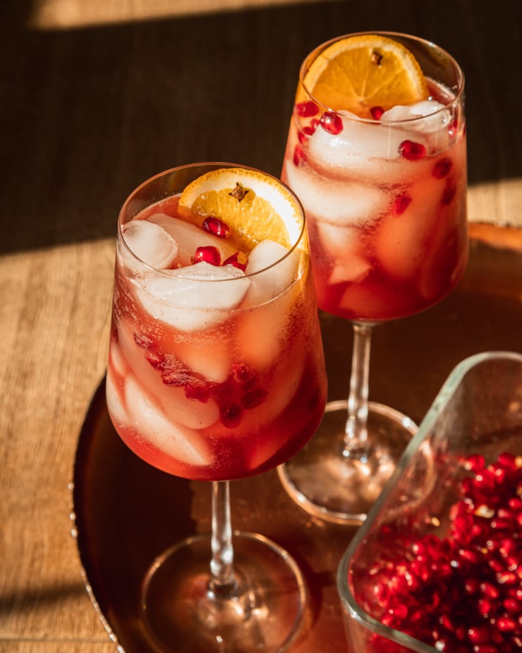 A 3/4 angle shot shows a pair of pomegranate pomander spritz mocktails in wine glasses. They are garnished with pomegranate seeds and slices of orange and set on top of a copper tray.