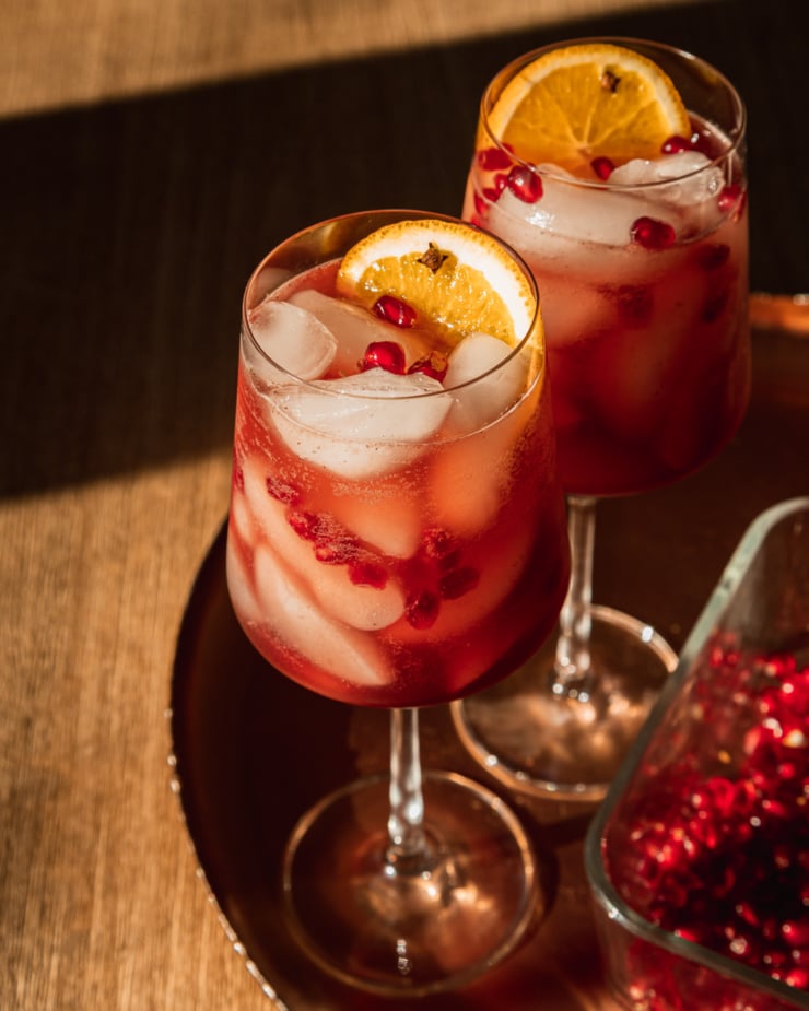 A 3/4 angle shot shows a pair of pomegranate pomander spritz mocktails in wine glasses. They are garnished with pomegranate seeds and slices of orange and set on top of a copper tray.