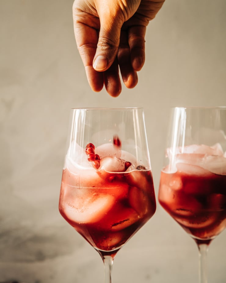 A head-on shot shows a hand garnishing a deep burgundy mocktail in a wine glass with pomegranate seeds/arils.