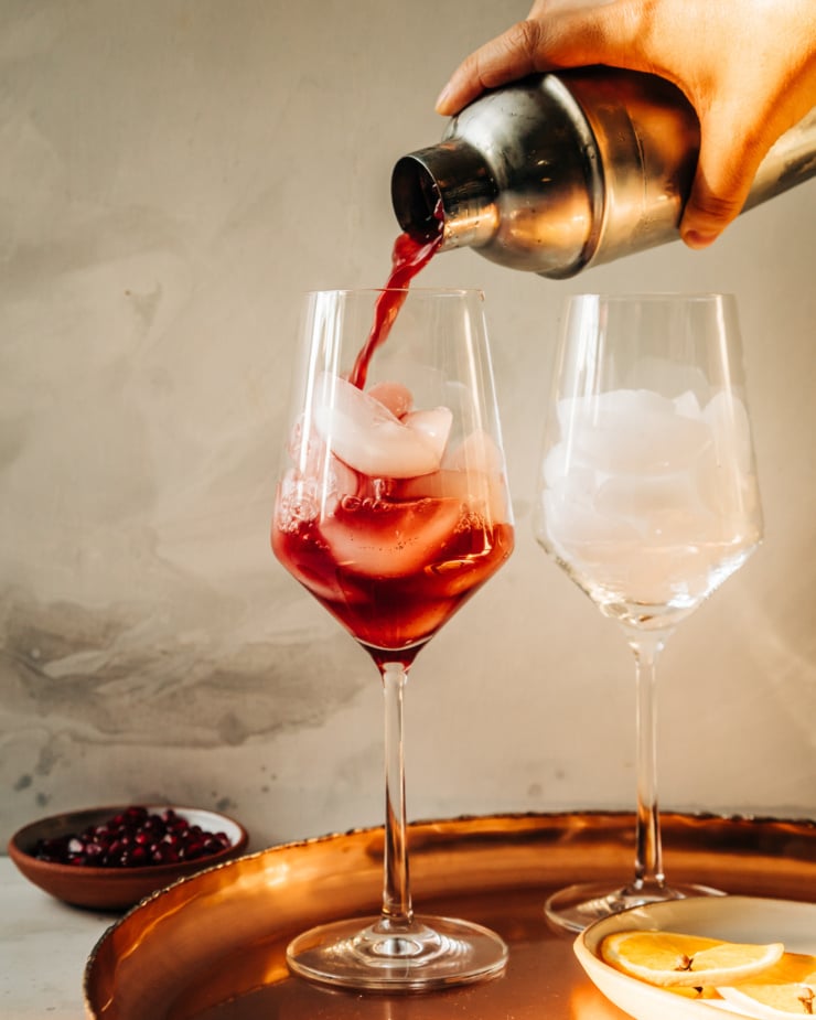 A head on shot shows a deep burgundy mocktail being strained and poured over ice in a wine glass.