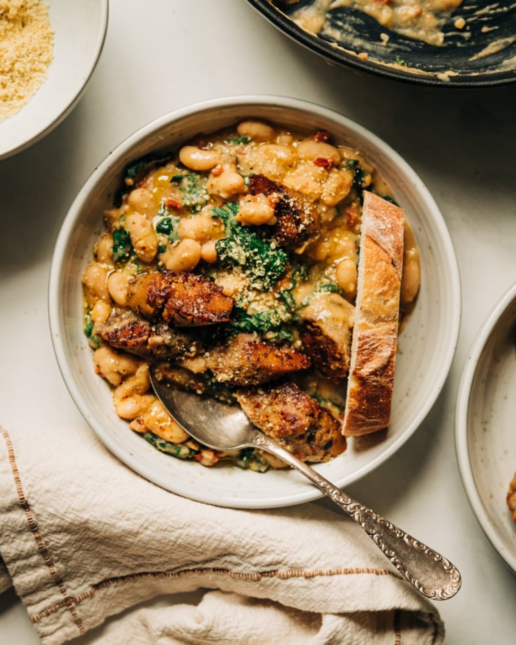 An overhead shot of a serving of a thick white bean and kale stew-like mixture in a shallow bowl. The stew is topped with browned slices of veggie sausage. A slice of crusty bread is perched in the bowl. An off-white linen napkin is seen to the side.