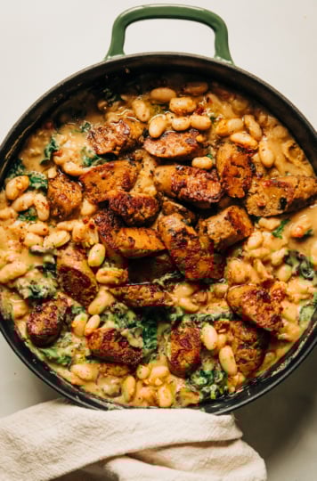 An overhead shot of a veggie sausage white bean skillet with lots of wilted kale. The skillet consists of a thick, stew-like mixture with browned slices of veggie sausage on top.