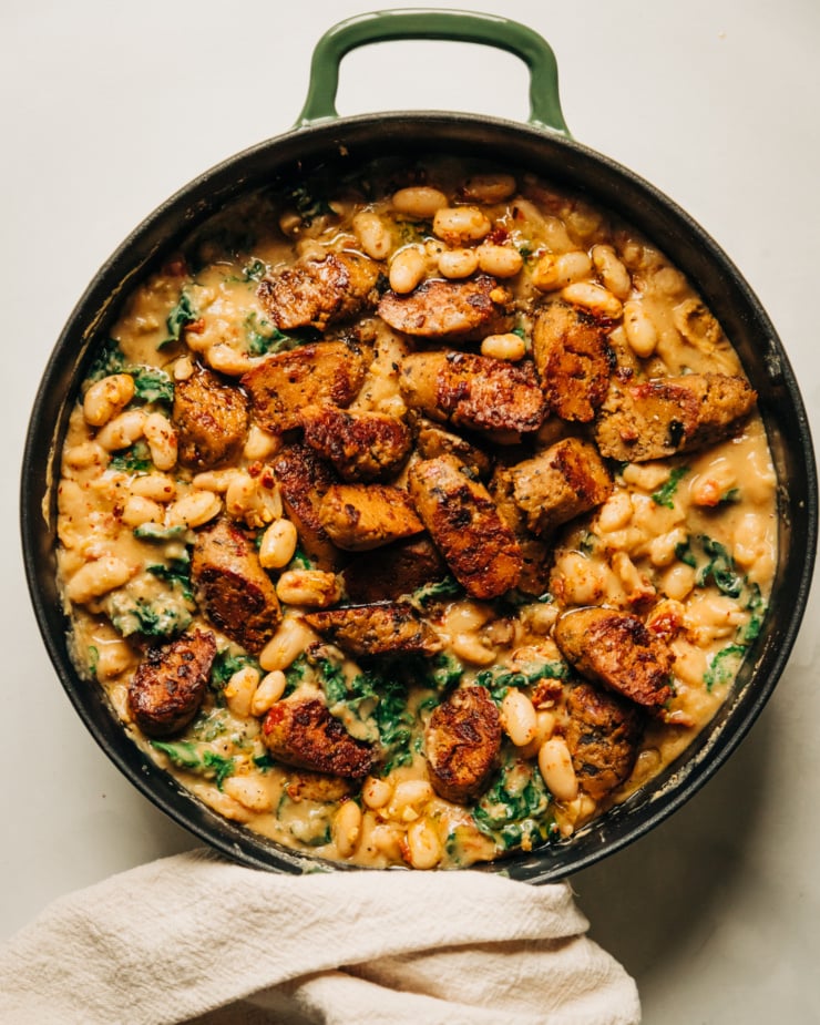 An overhead shot of a veggie sausage white bean skillet with lots of wilted kale. The skillet consists of a thick, stew-like mixture with browned slices of veggie sausage on top.