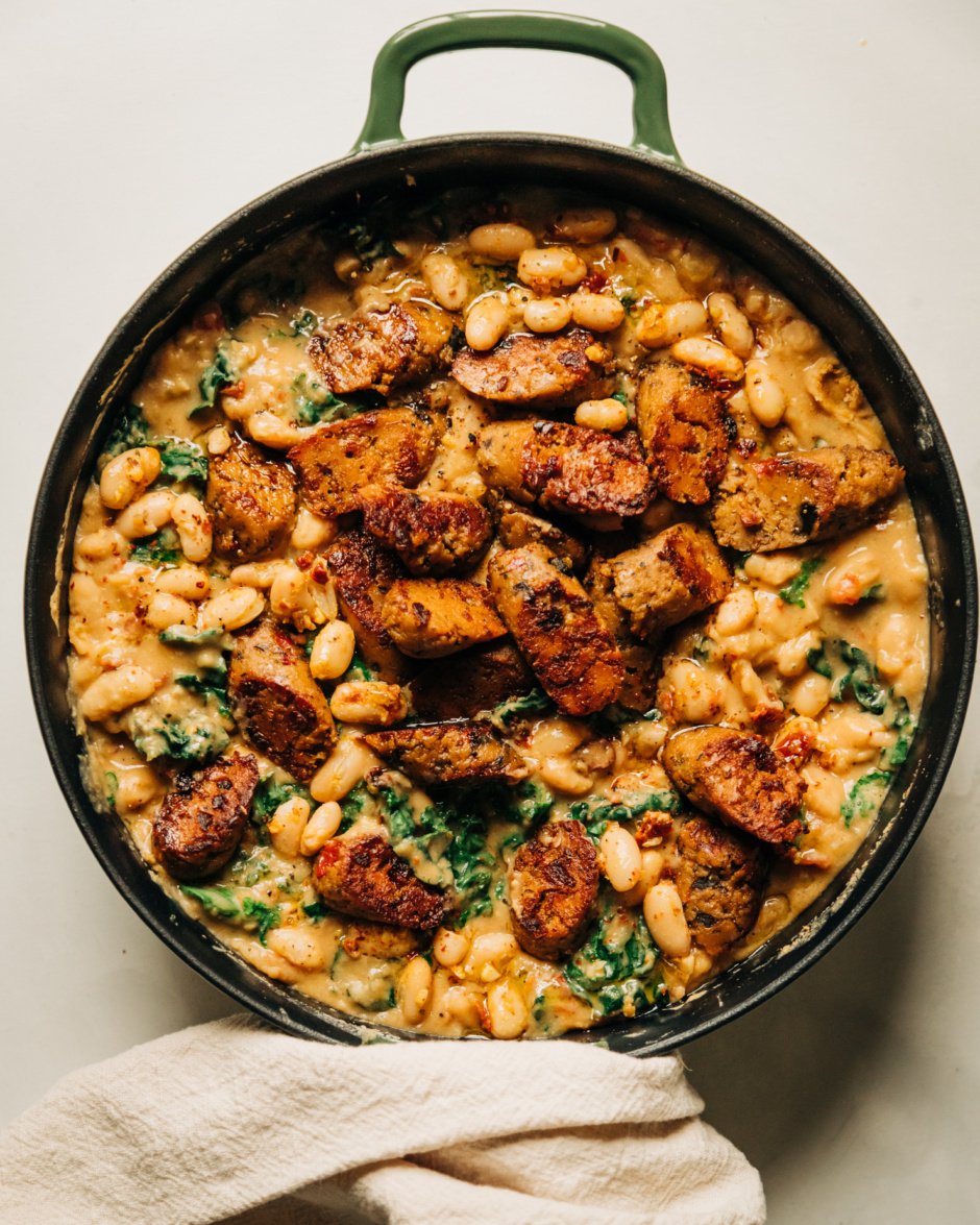 An overhead shot of a veggie sausage white bean skillet with lots of wilted kale. The skillet consists of a thick, stew-like mixture with browned slices of veggie sausage on top.