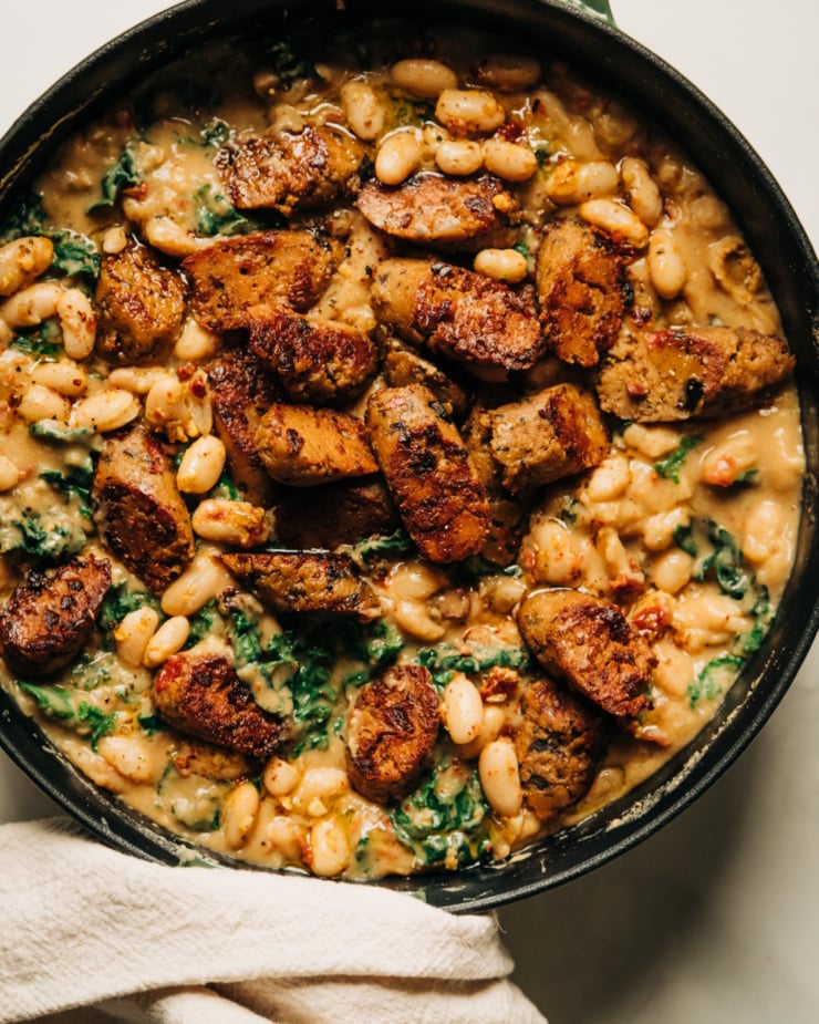 An overhead shot of a veggie sausage white bean skillet with lots of wilted kale. The skillet consists of a thick, stew-like mixture with browned slices of veggie sausage on top. The skillet is garnished with ground chillies.