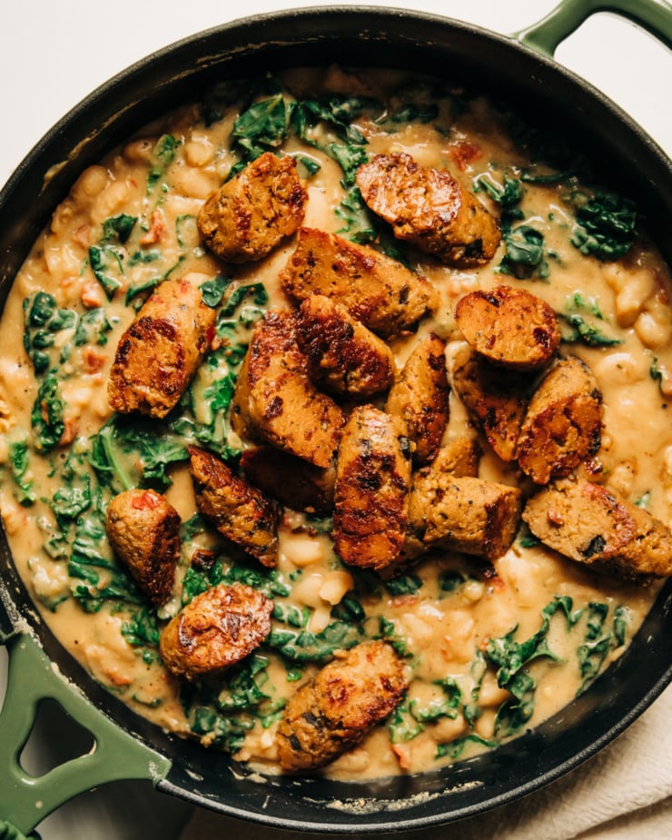 An overhead shot of a veggie sausage white bean skillet with lots of wilted kale. The skillet consists of a thick, stew-like mixture with browned slices of veggie sausage on top.