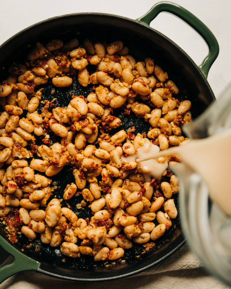 An overhead shot of a creamy mixture being poured into a skillet with cooked white beans, sun dried tomatoes, and spices.