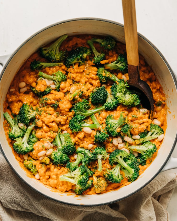 An overhead shot of cheesy vegan broccoli and barley with white beans in a Dutch oven-style pot. A wooden handle is sticking out of the pot.