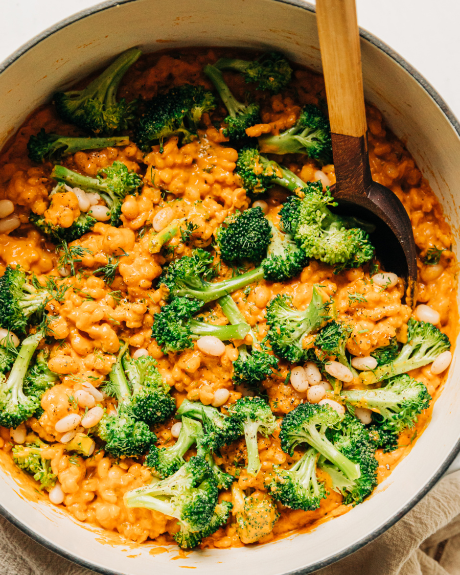 An up close, overhead shot of cheesy vegan broccoli and barley with white beans in a Dutch oven-style pot. A wooden handle is sticking out of the pot.
