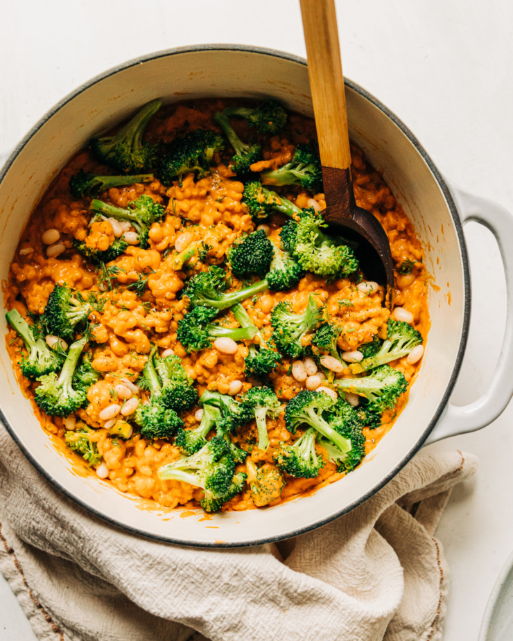 An overhead shot of cheesy vegan broccoli and barley with white beans in a Dutch oven-style pot. A wooden handle is sticking out of the pot.